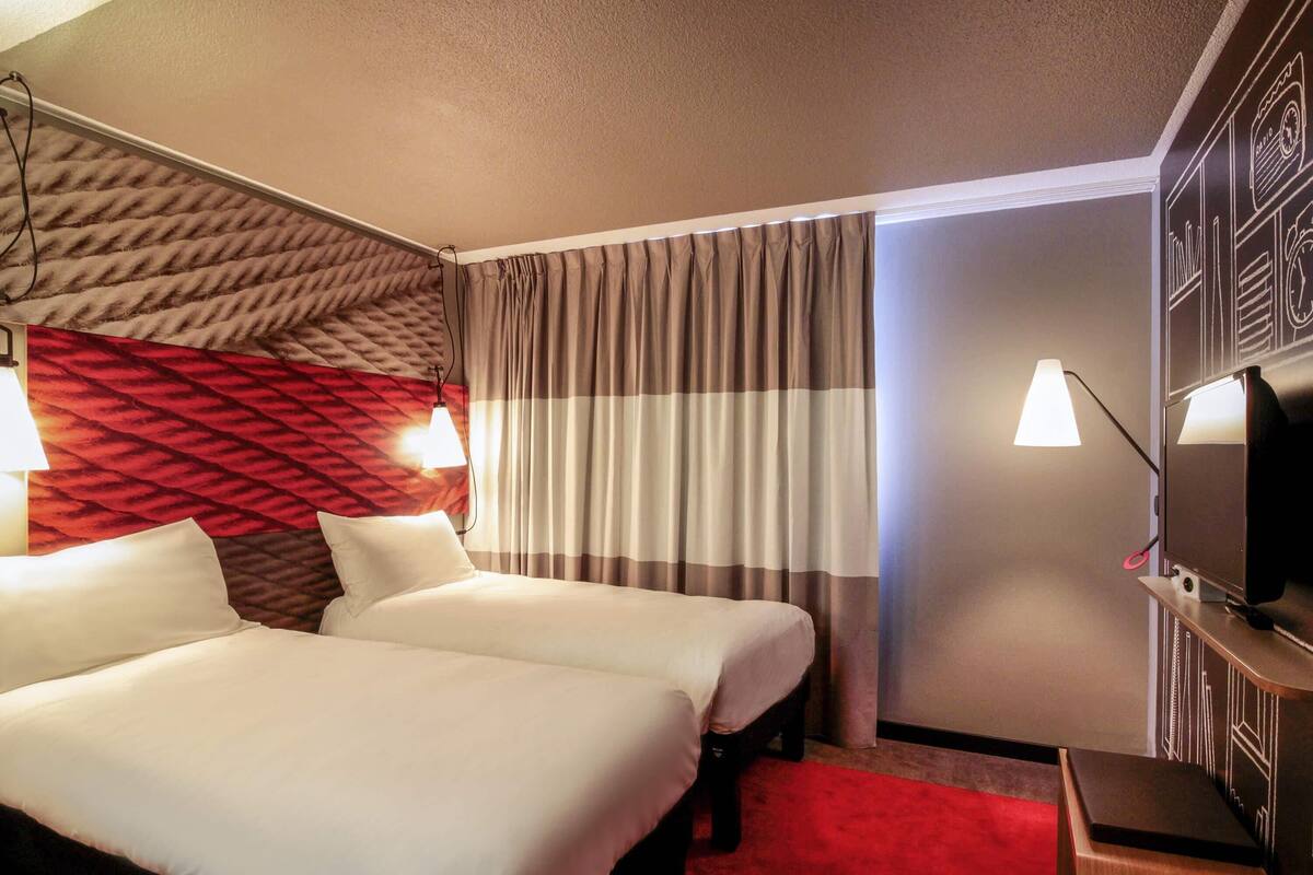 A hotel room with two beds, a red carpet, a television on a stand, and a wall with a geometric pattern.
