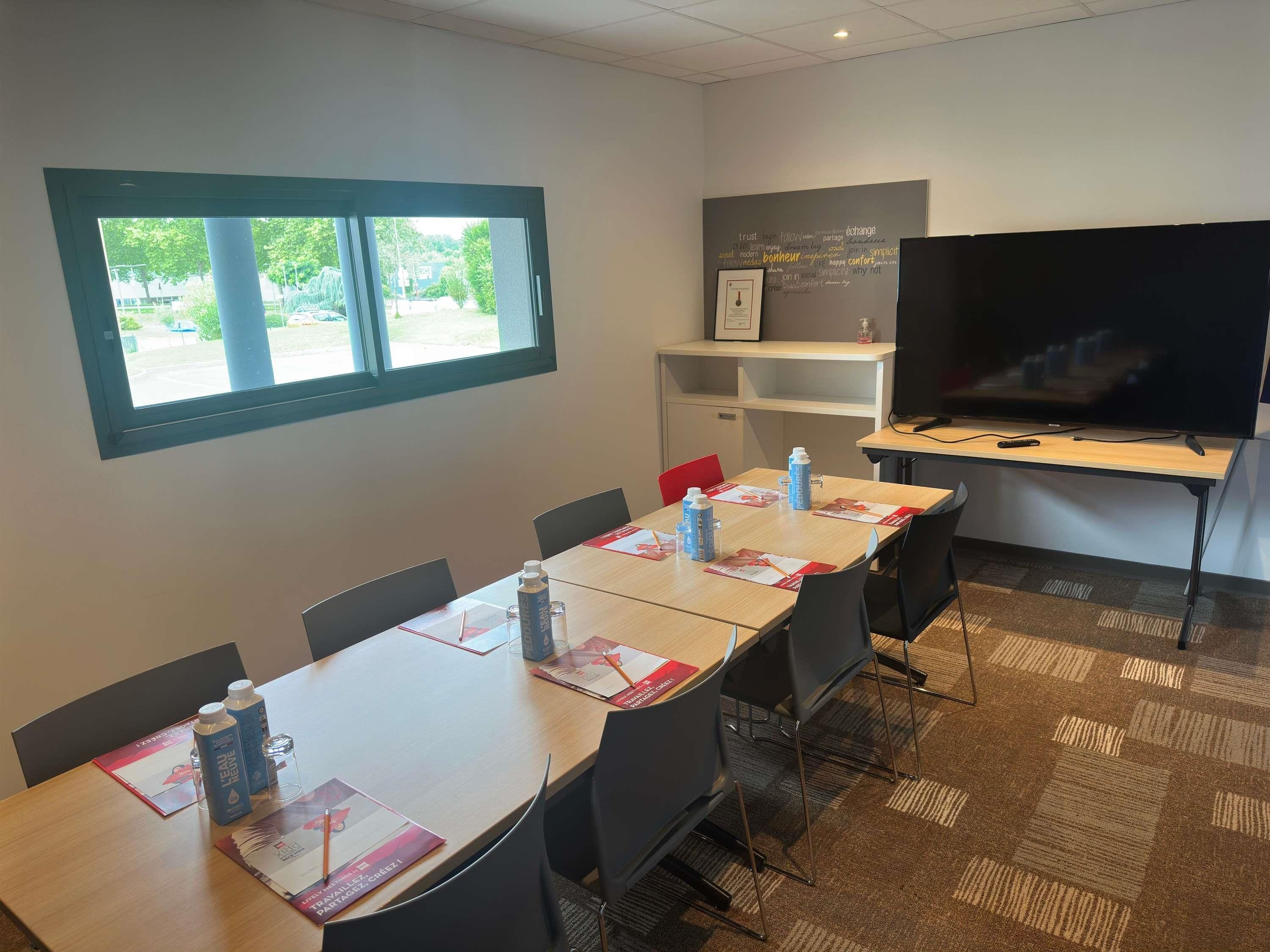 A conference room with a long table, chairs, a projector screen, a television, and a window with a view of greenery.