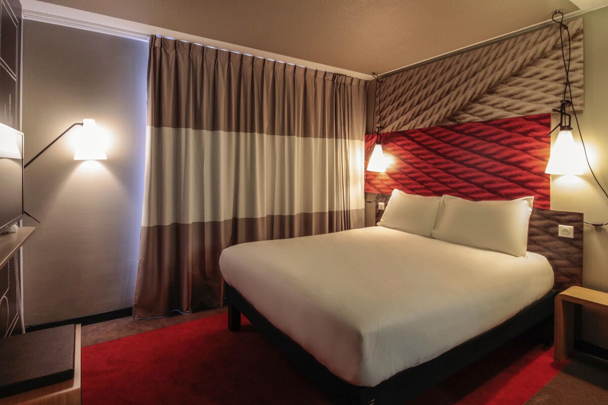 A hotel room with a bed, curtains, a red carpet, and wall-mounted lamps.