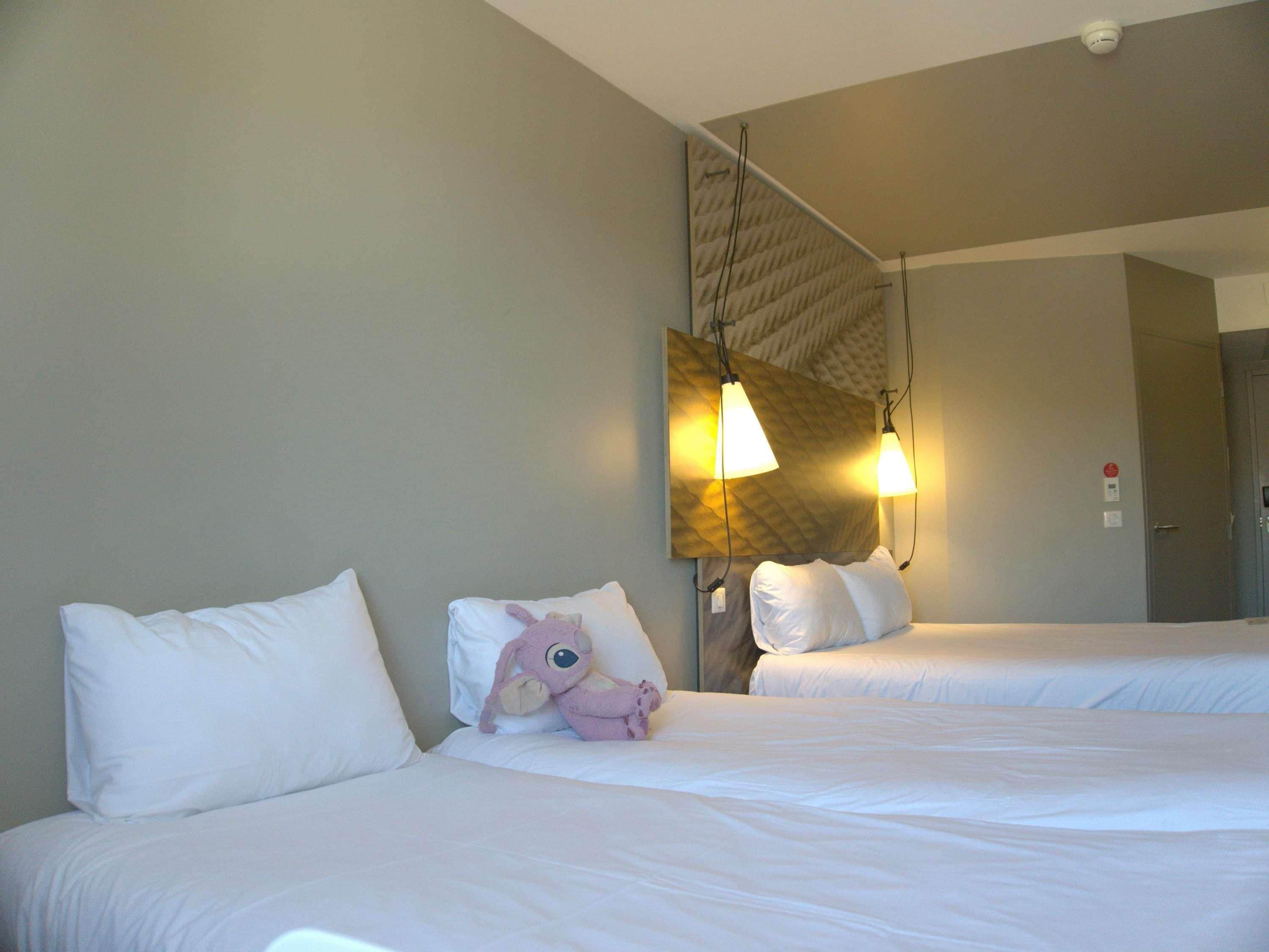 A hotel room with two beds, white bedding, a plush toy on one bed, and a wall-mounted light fixture.