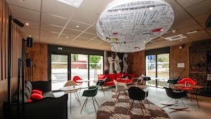 A modern interior with a central seating area, a large wall-mounted light fixture, and a view of the outdoors through large windows. - ibis Cahors (Cahors)