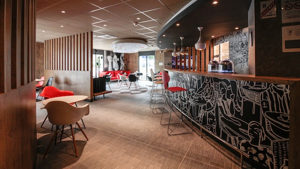 A modern bar area with a mural, wooden paneling, and red chairs. - ibis Cahors (Cahors)