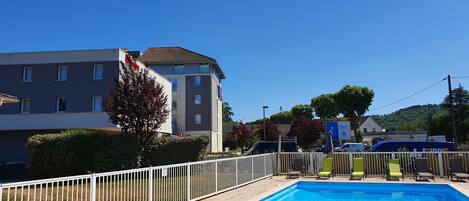 Une piscine entourée d’une clôture blanche, un bâtiment doté de nombreuses fenêtres et un ciel d’un bleu limpide.