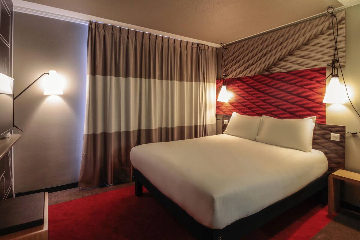 A hotel room with a bed, curtains, a red carpet, and wall-mounted lamps.