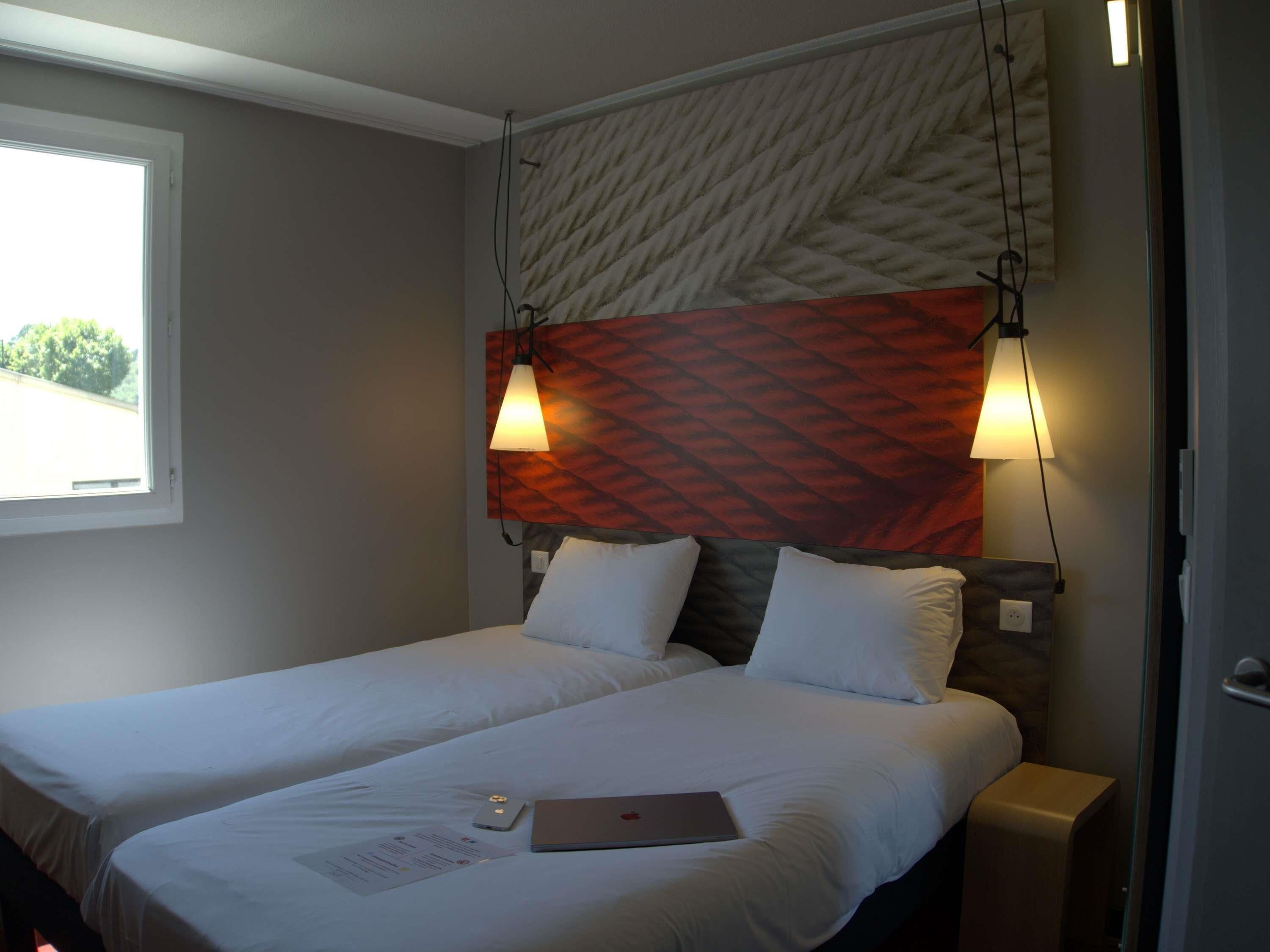 Une chambre d’hôtel avec deux lits, une fenêtre et une lampe fixée au mur.