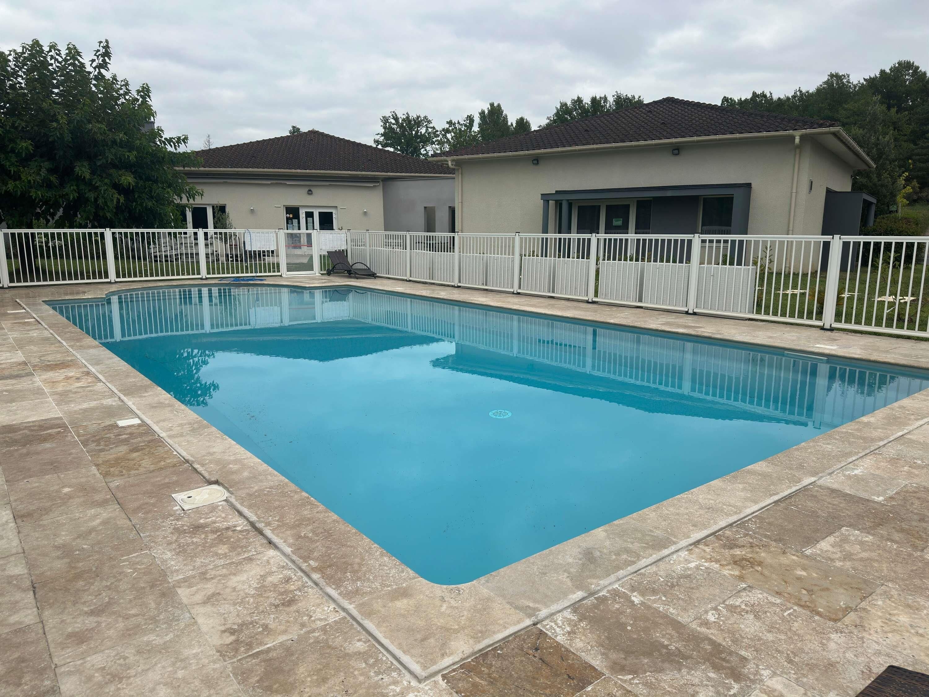 A rectangular swimming pool with a tiled deck, surrounded by a white fence.