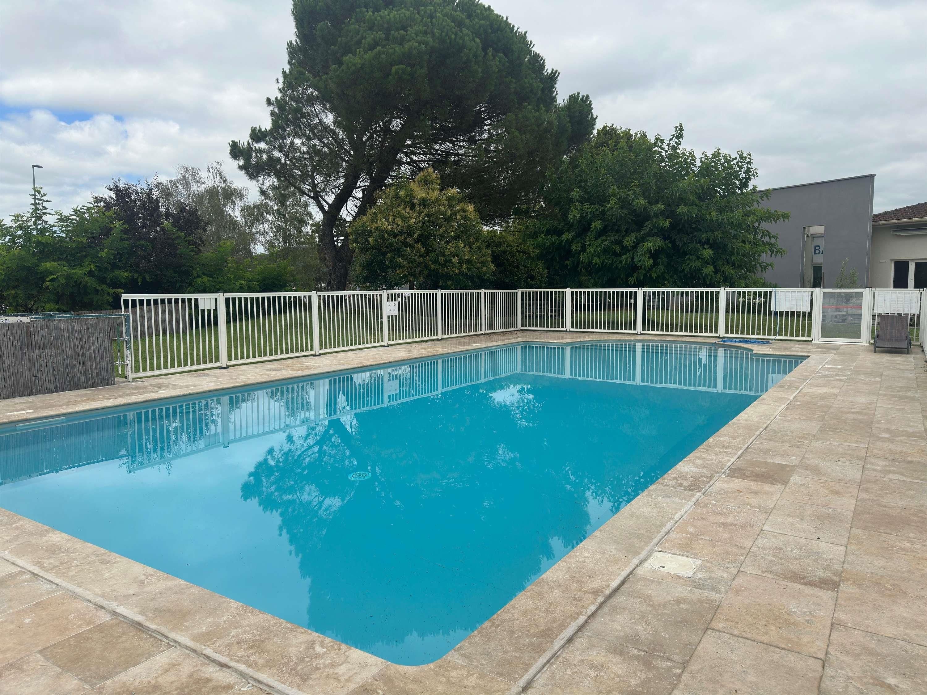 A rectangular swimming pool with a white fence surrounding it, set against a backdrop of trees and a building.