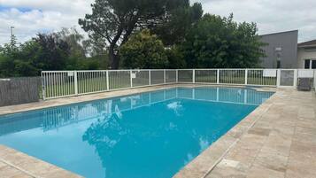 A rectangular swimming pool with a white fence surrounding it, set against a backdrop of trees and a building.