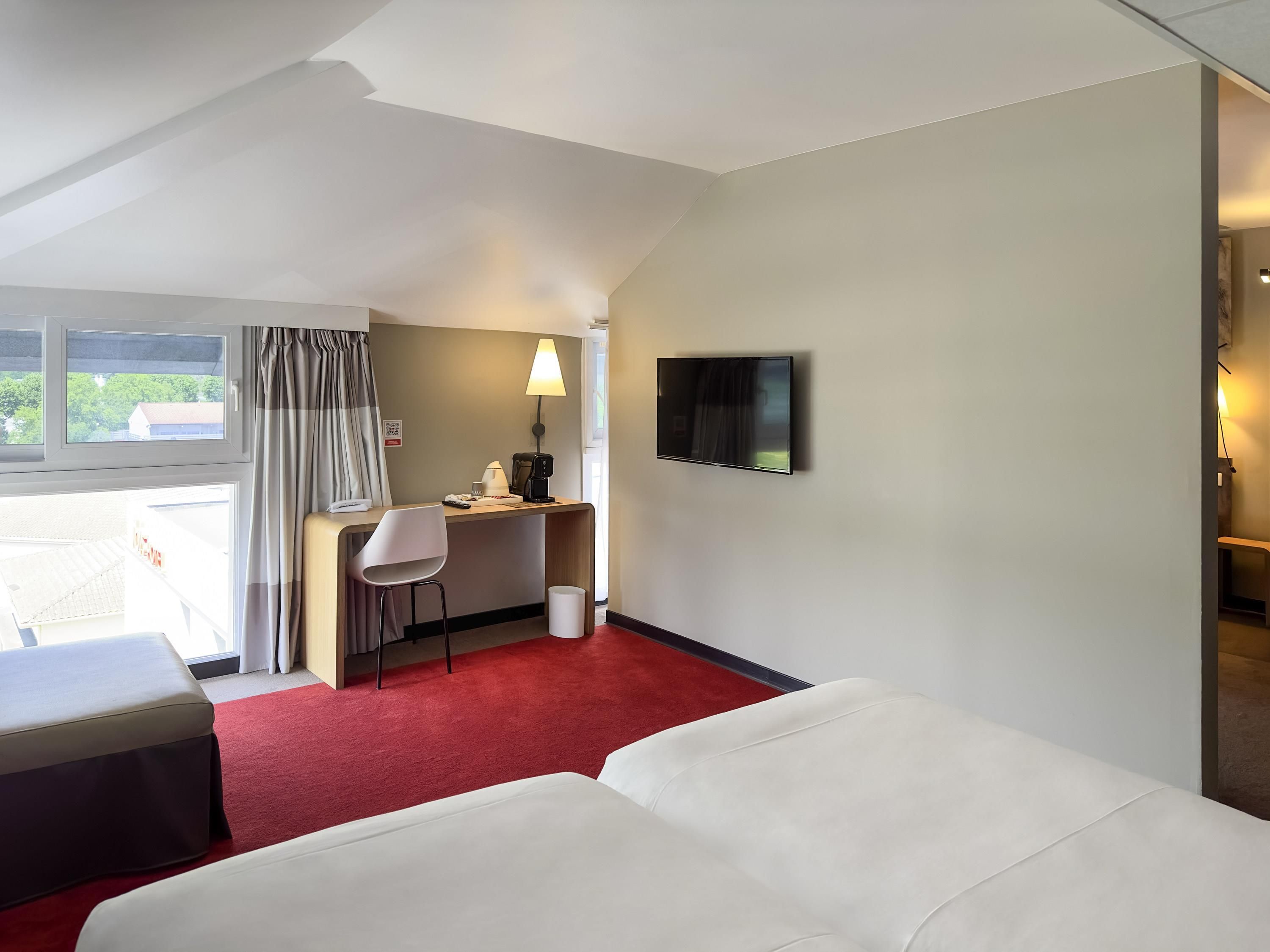Une chambre d’hôtel comprenant un lit, un bureau avec une chaise, un téléviseur fixé au mur et une fenêtre avec des rideaux.