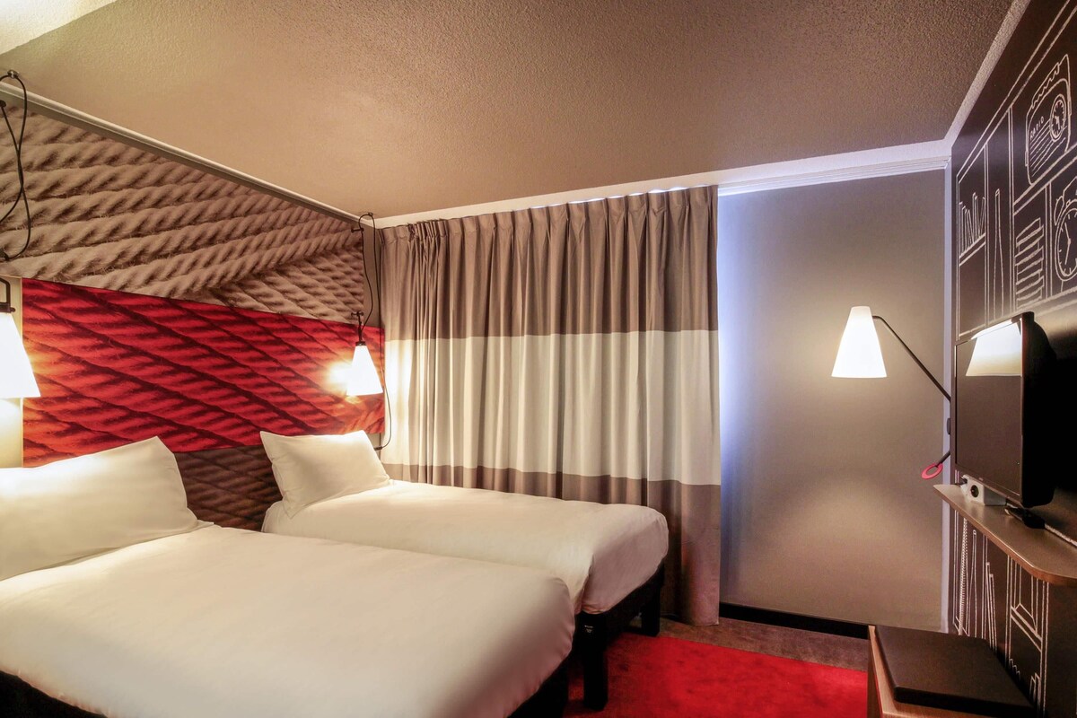 A hotel room with two beds, a red carpet, a television on a stand, and a wall with a geometric pattern.