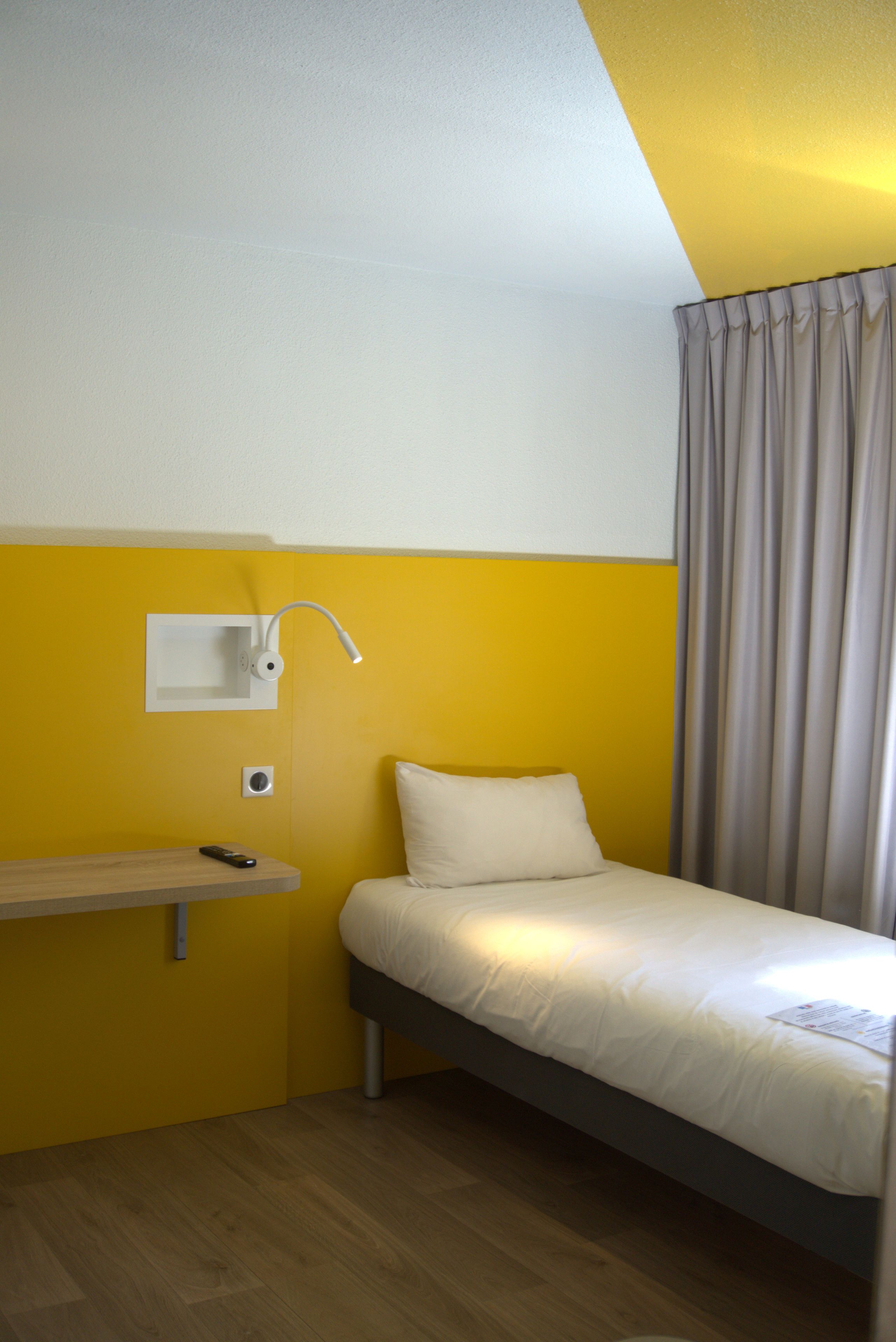 A small, single-bed room with a wooden floor, a bedside table, a lamp, and a curtain.