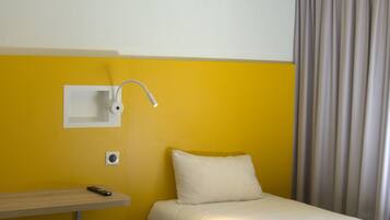 A small, single-bed room with a wooden floor, a bedside table, a lamp, and a curtain.
