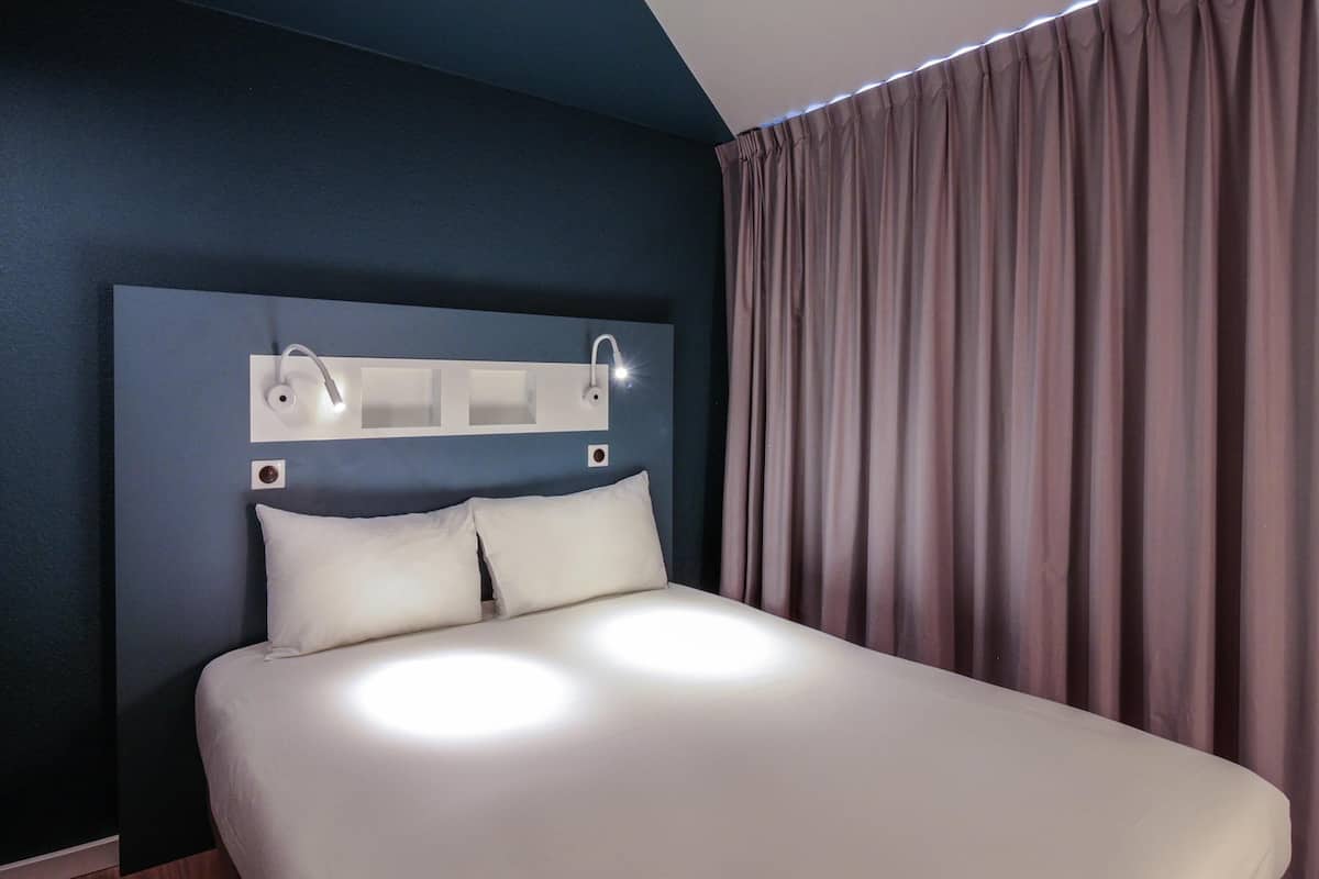 Ein Hotelzimmer mit einem großen Bett, einem Kopfteil mit integrierter Beleuchtung und einem Fenster mit Vorhängen.