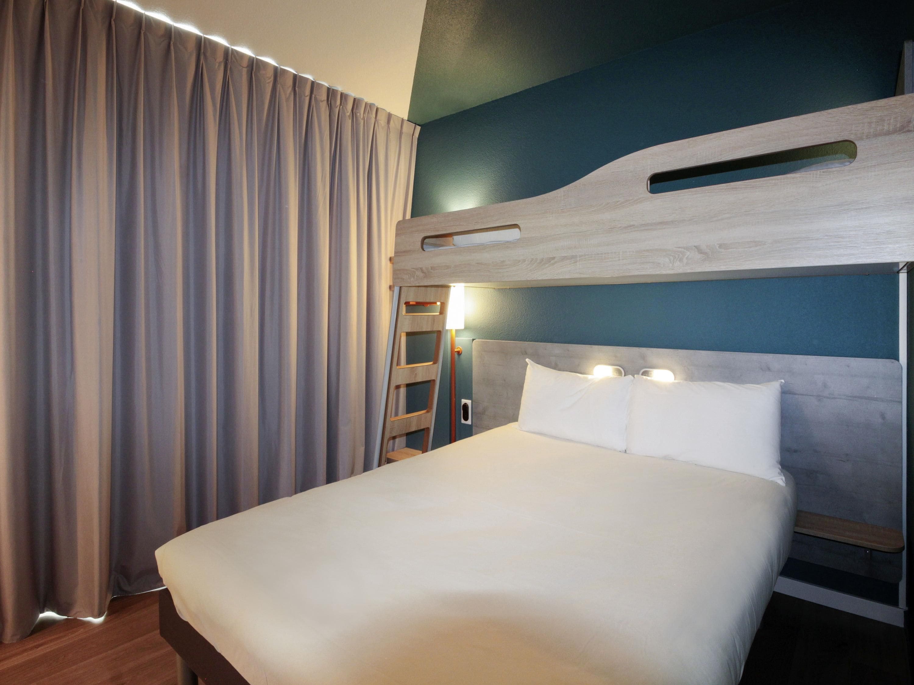 Una habitación de hotel con literas, un cabecero de madera, un ventanal con cortinas y una mesita de noche con lámpara.