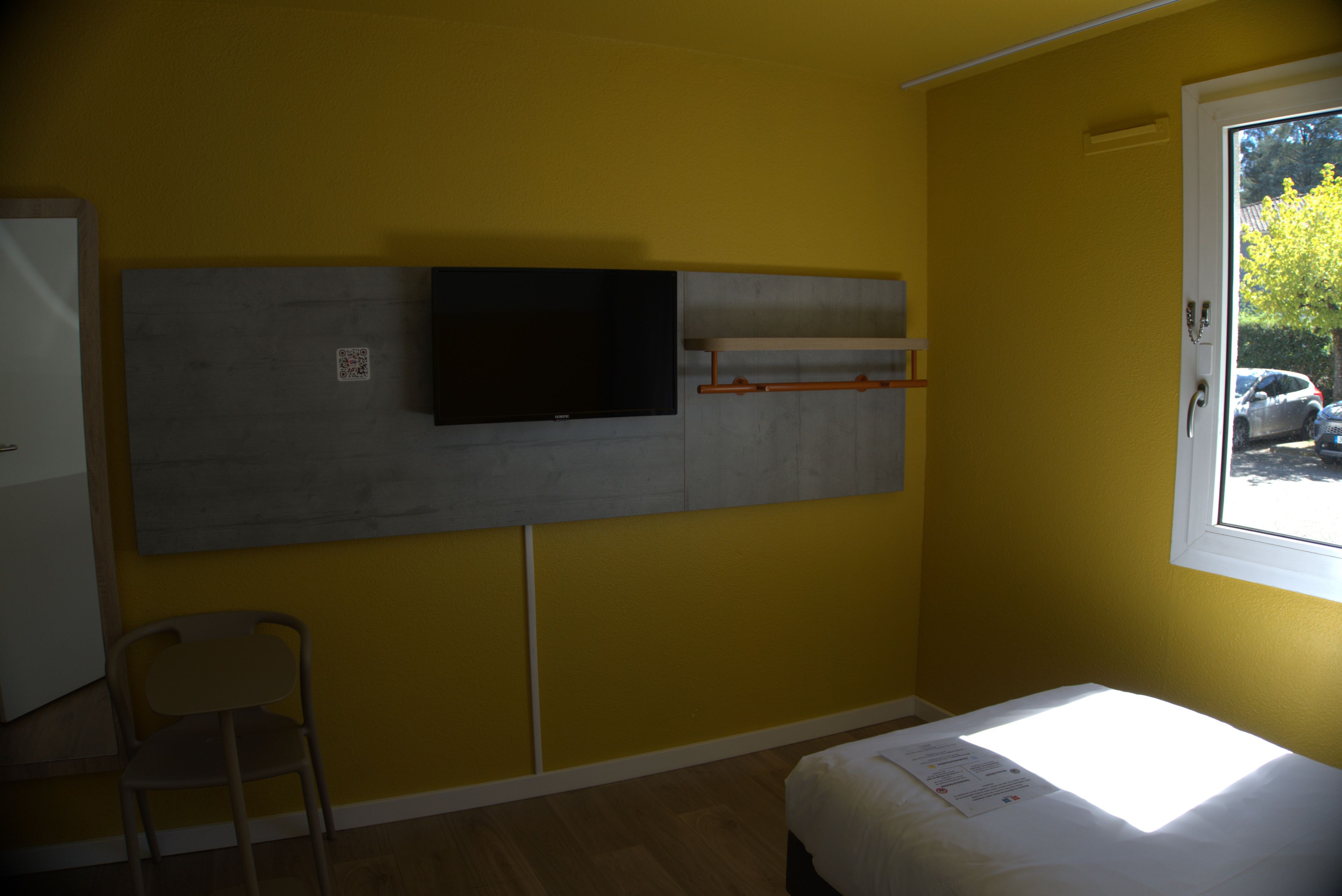 Een kamer met een bed, een stoel, een televisie aan de muur en een raam met uitzicht.