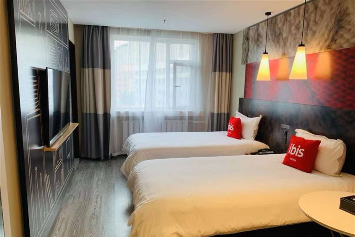 A hotel room with two beds, a TV, a small table, and a view of the outside through large windows.