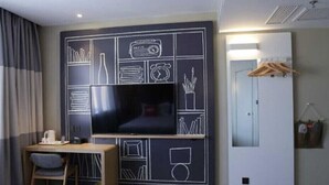 A modern hotel room with a large flat-screen TV mounted on a black wall with white geometric designs. There's a wooden desk with a chair and a small trash bin. A mirror and a wooden hanger rack are visible on the right side. - Ibis Jilin Beishan Park Hotel (Jilin)