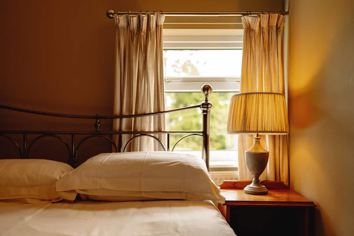 A neatly made bed with white linens, a bedside table with a lamp, and a window with curtains.