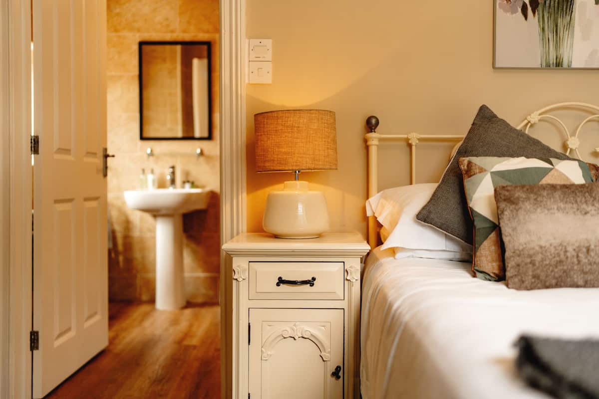 A bedroom with a bed, a nightstand with a lamp, a bedside table, and a wall-mounted mirror.
