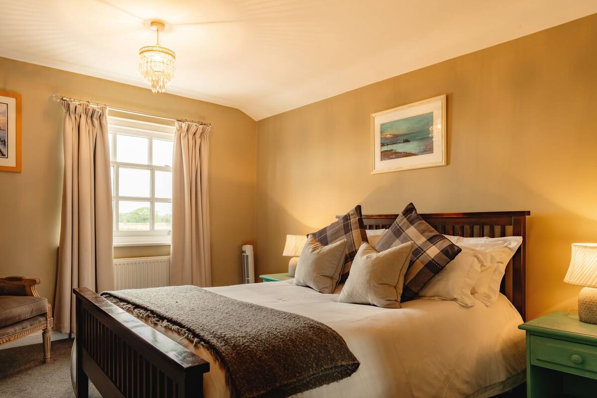 A neatly arranged bedroom with a large bed, a bedside table, a chair, and a window with curtains.