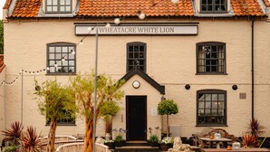 A building with a sign reading "Wheatacre White Lion." It features a front entrance with a door, a small porch area with seating, and potted plants around. - Wheatacre White Lion (Beccles)