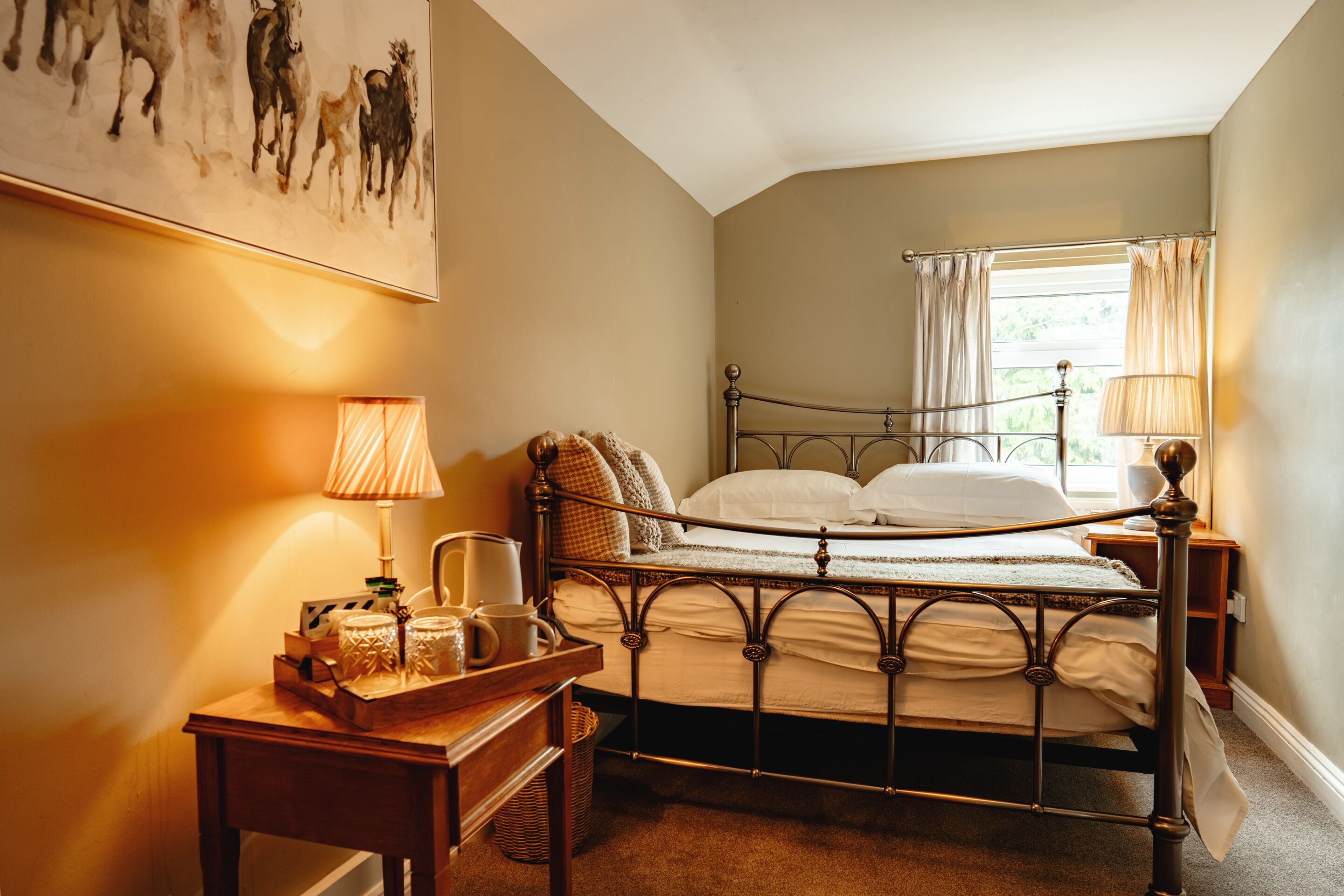 A bedroom with a metal bed frame, a bedside table with a lamp, a window with curtains, and a painting on the wall.