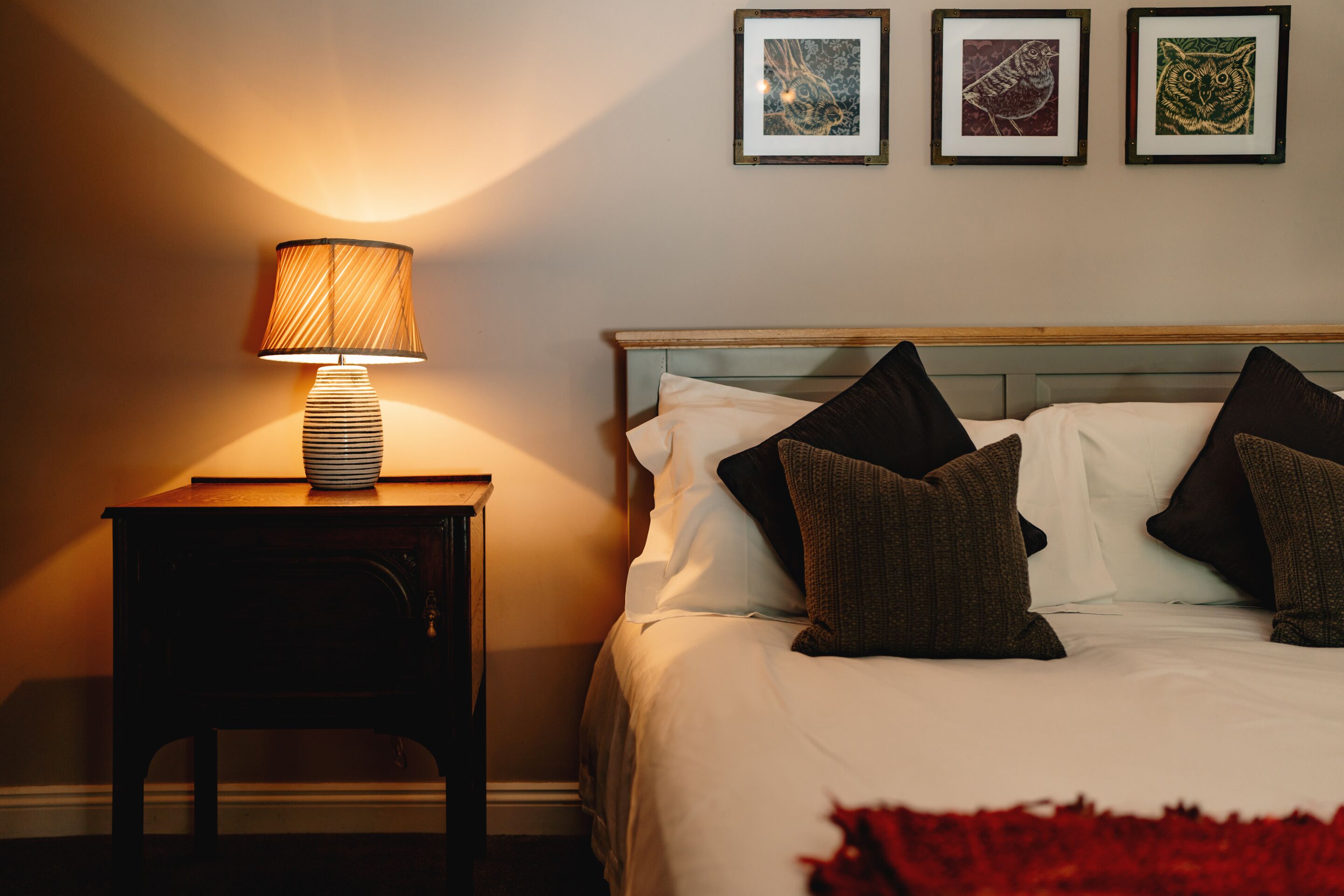 A neatly made bed with white linens and dark pillows, a bedside table with a lamp, and three framed pictures on the wall.