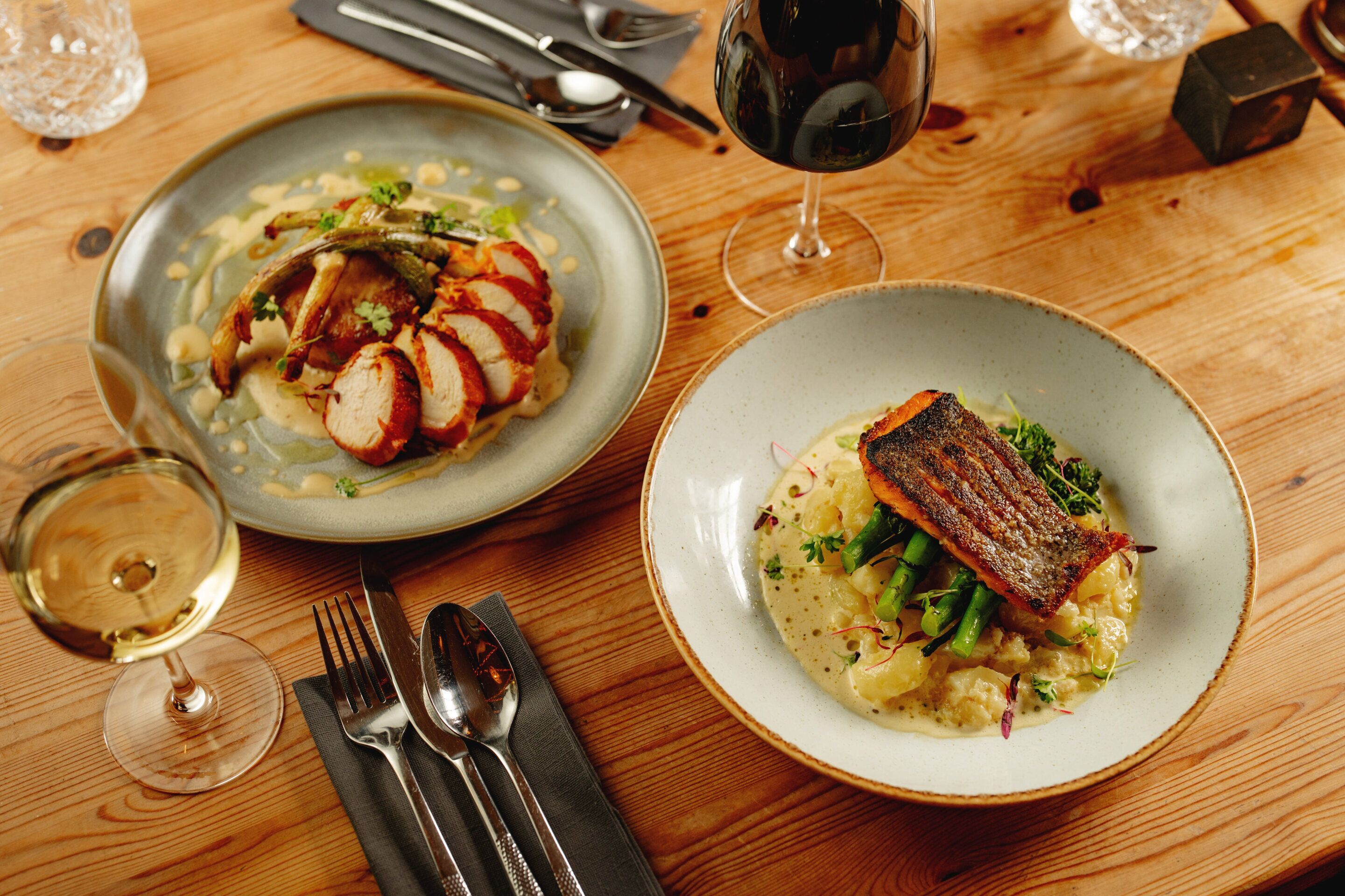 Two plates of food on a wooden table with cutlery and glasses.