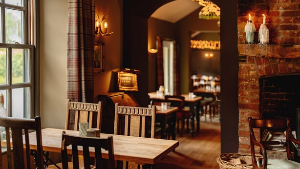 A cozy restaurant interior with wooden tables and chairs, a brick fireplace, and a view through an arched doorway. - Wheatacre White Lion (Beccles)