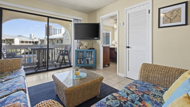 A living room with wicker furniture, a flat-screen TV, and a view of buildings through a glass door. - Las Palmas 224 2 Bedroom Condo by RedAwning (Gulf Shores)
