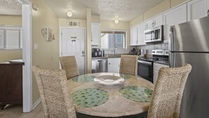 A modern kitchen with white cabinets, a stainless steel refrigerator, and a round table with woven chairs. - Las Palmas 224 2 Bedroom Condo by RedAwning (Gulf Shores)