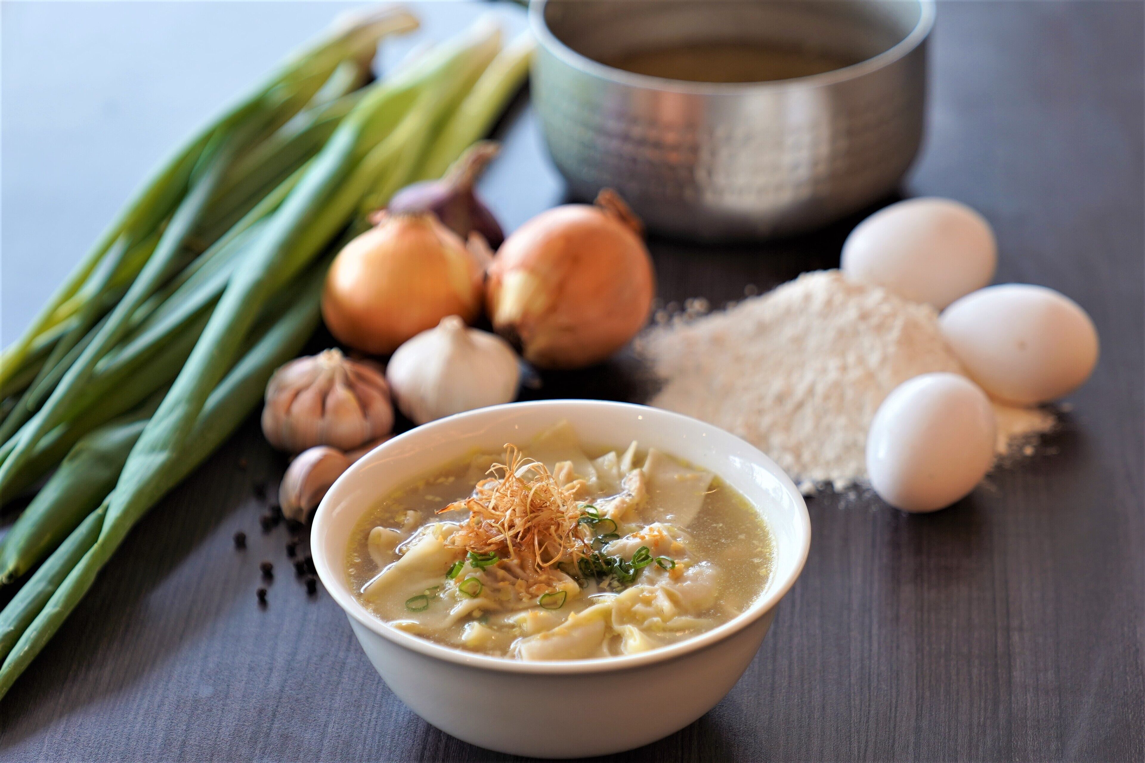 A bowl of soup with noodles, garnished with green onions and fried shallots, surrounded by ingredients like green onions, garlic, eggs, and a bowl of broth.