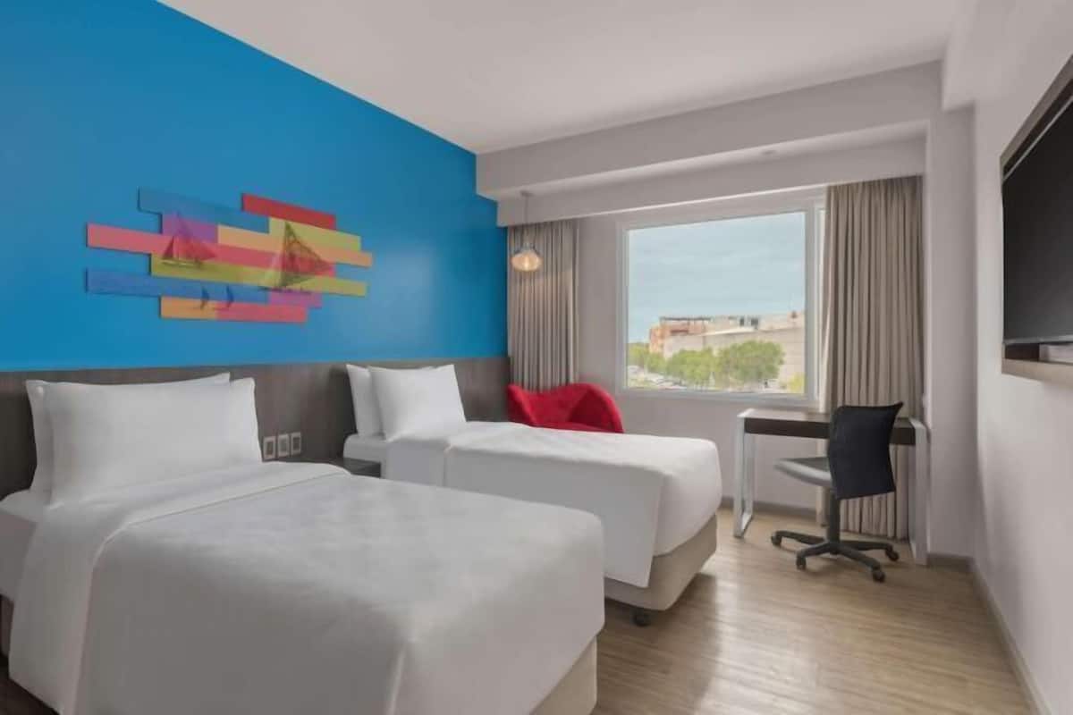 Ein Hotelzimmer mit zwei Betten, einem Schreibtisch, einem Sessel, einem Fenster mit Ausblick und einer farbenfrohen Wanddekoration.