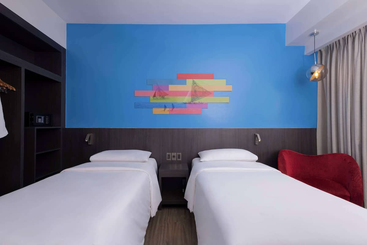 Ein Hotelzimmer mit zwei Betten, einer blauen Akzentwand, einem roten Sessel und einer modernen Leuchte.