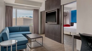 A modern hotel room with a blue sofa, a small table, a TV mounted on the wall, and a bed with a red pillow.