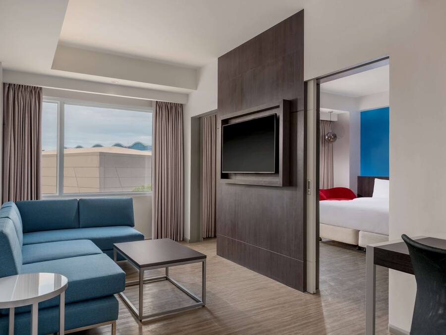 A modern hotel room with a blue sofa, a small table, a TV mounted on the wall, and a bed with a red pillow.