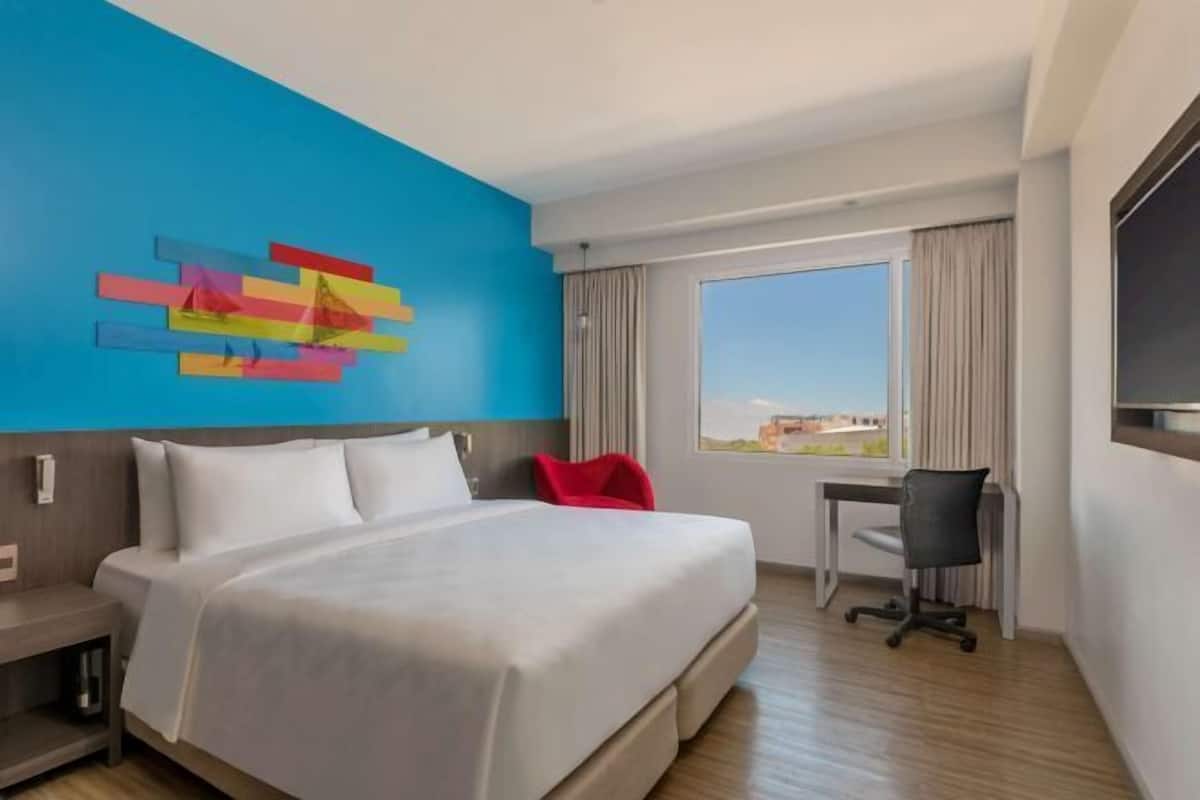 Ein Hotelzimmer mit Bett, Schreibtisch, Stuhl, Fenster mit Aussicht und einer farbenfrohen Wanddekoration.