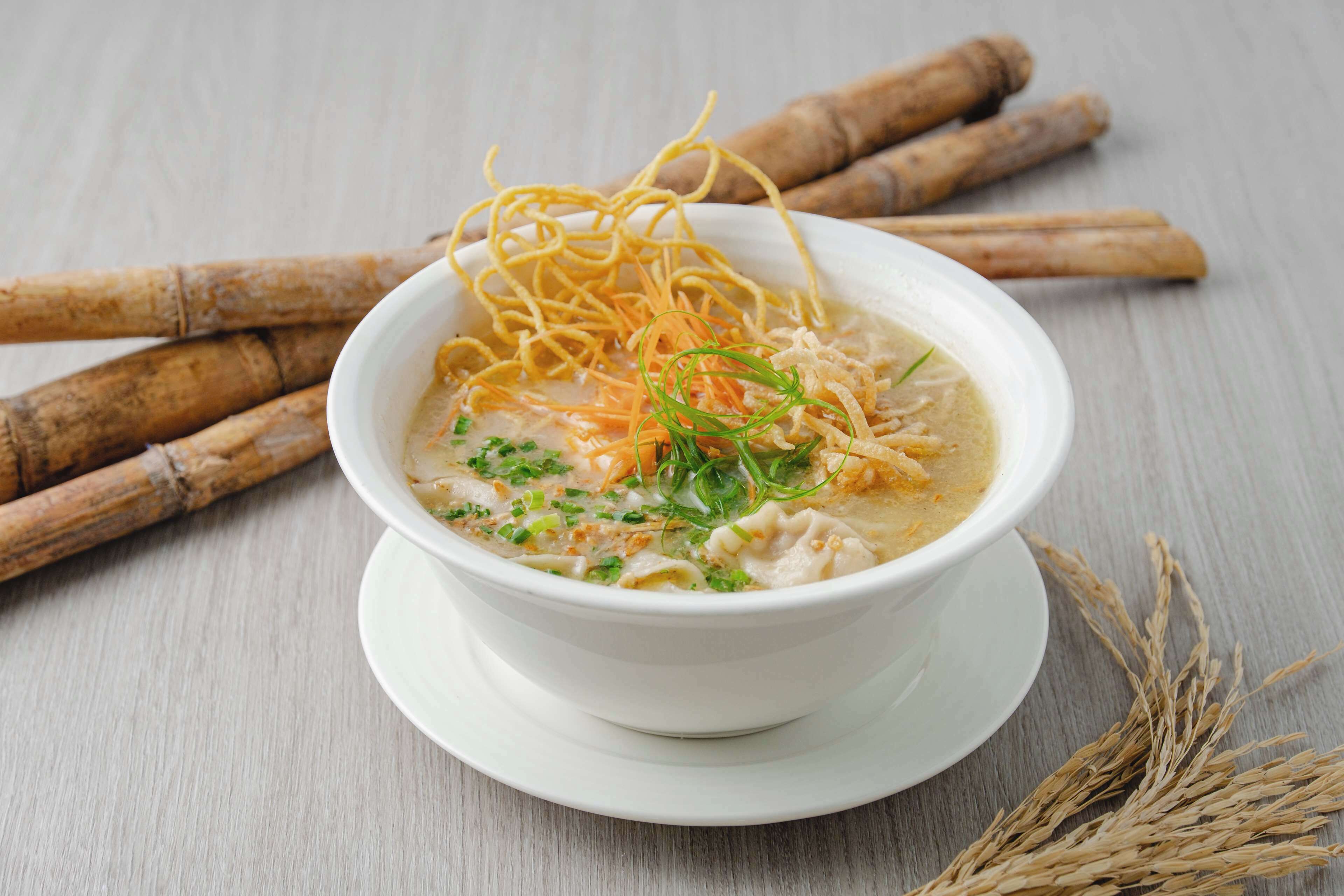 Un tazón de sopa con fideos, decorado con hierbas y vegetales rallados, sobre un plato blanco y su platillo.