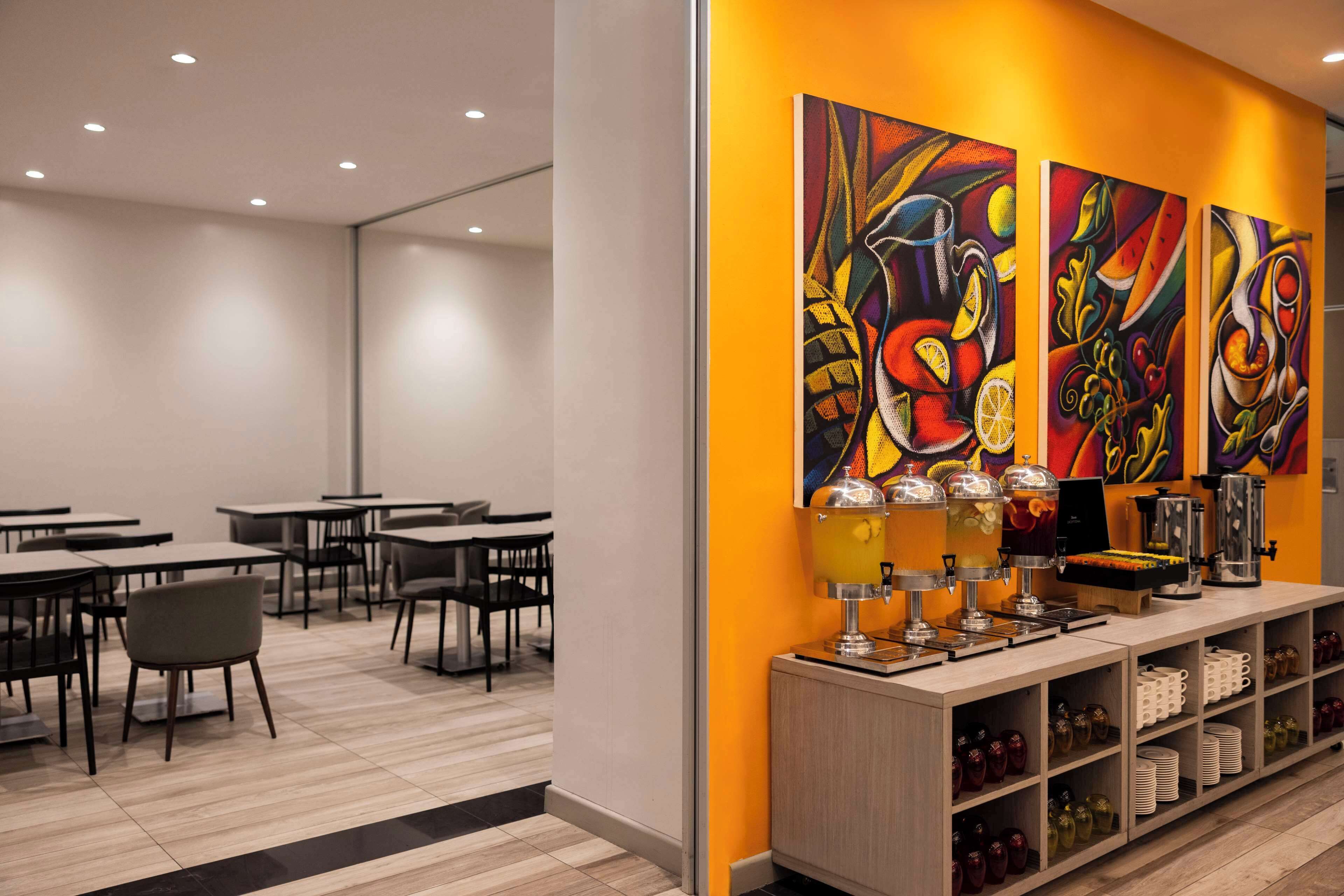 Una cafeteria moderna amb taules i cadires, una zona de begudes i obra d'art colorida a la paret.