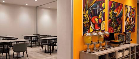 A modern cafe with tables and chairs, a beverage station, and colorful artwork on the wall.