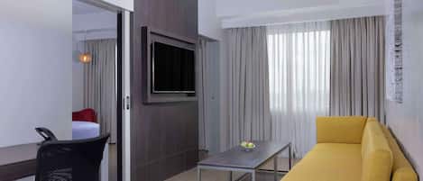 A modern hotel room with a sofa, a small table, a TV mounted on a wooden panel, and a chair.