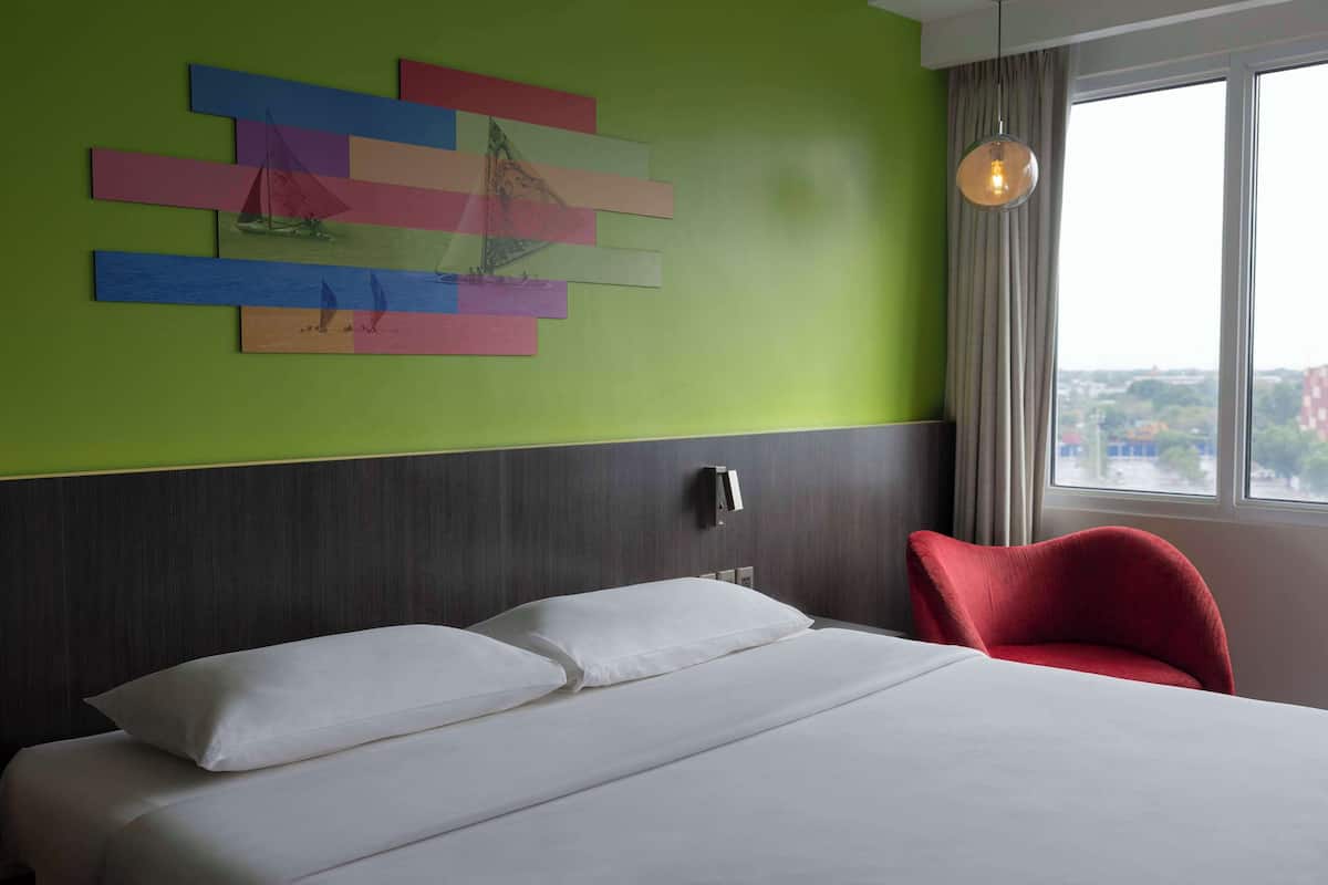 Ein Hotelzimmer mit Bett, einem roten Sessel, einer Wand mit farbenfroher Kunst, einem Fenster mit Blick und einer Pendelleuchte.