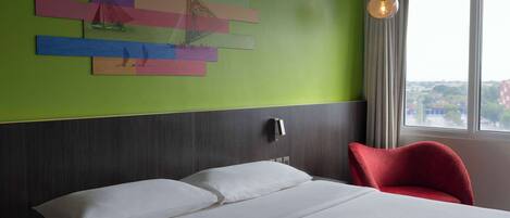 A hotel room with a bed, a red chair, a wall with colorful artwork, a window with a view, and a hanging light fixture.