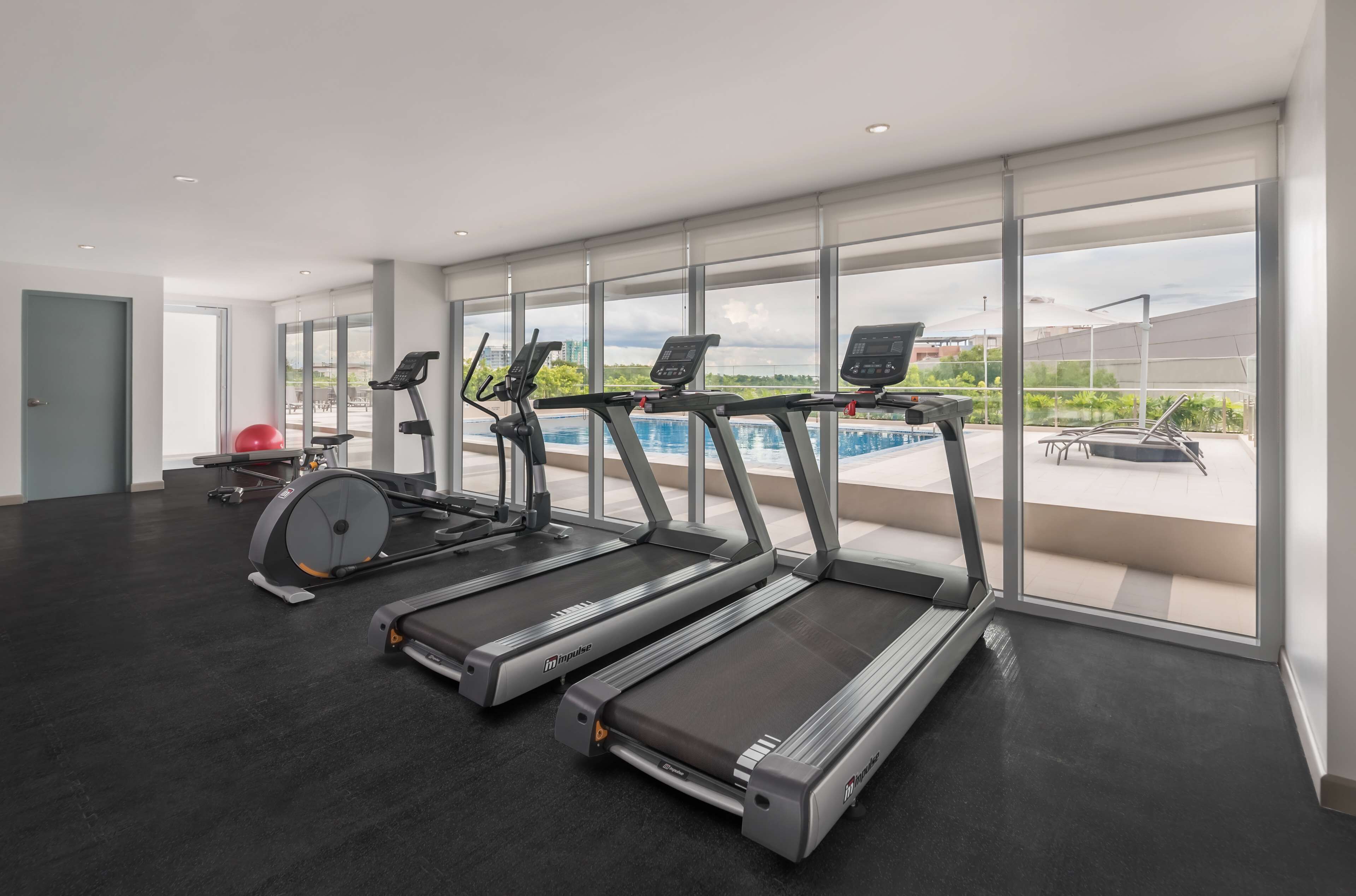 A modern gym with treadmills, a rowing machine, and a red exercise ball. Large windows offer views of a swimming pool and greenery.