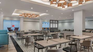 A modern dining area with tables and chairs, a kitchen in the background, and a bright ceiling with pendant lights.