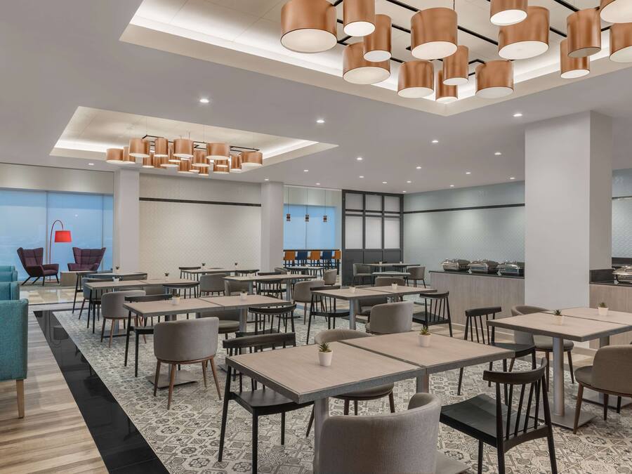 A modern dining area with tables and chairs, a kitchen in the background, and a bright ceiling with pendant lights.