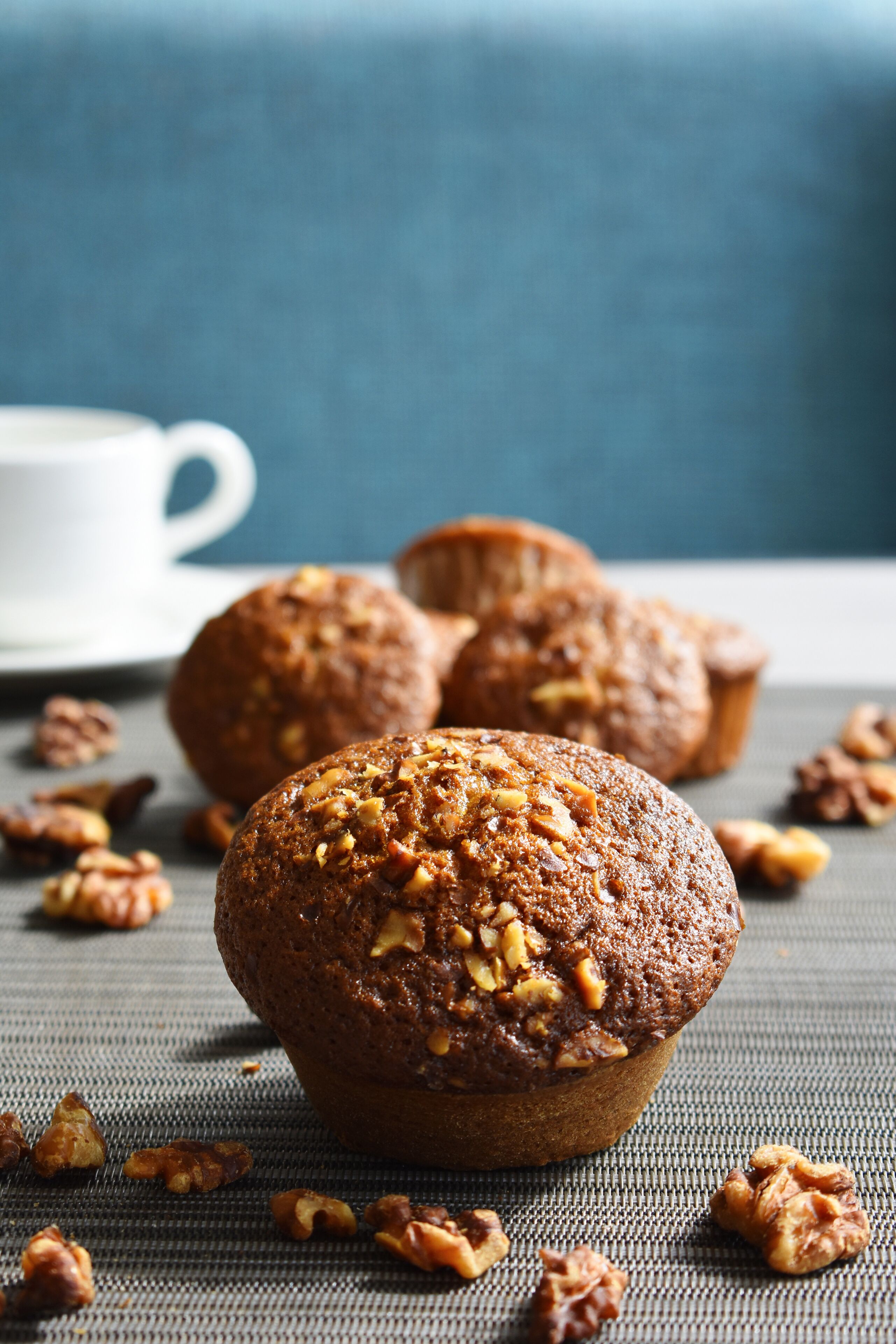 Trois muffins aux noix sur une surface texturée, une tasse blanche en arrière-plan et des noix éparpillées autour.