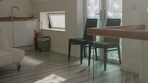 A spacious room with a sloped ceiling, a wooden floor, a dining table with chairs, a white sofa, a small kitchen area with a sink and a fire extinguisher, and a window with a butterfly curtain.