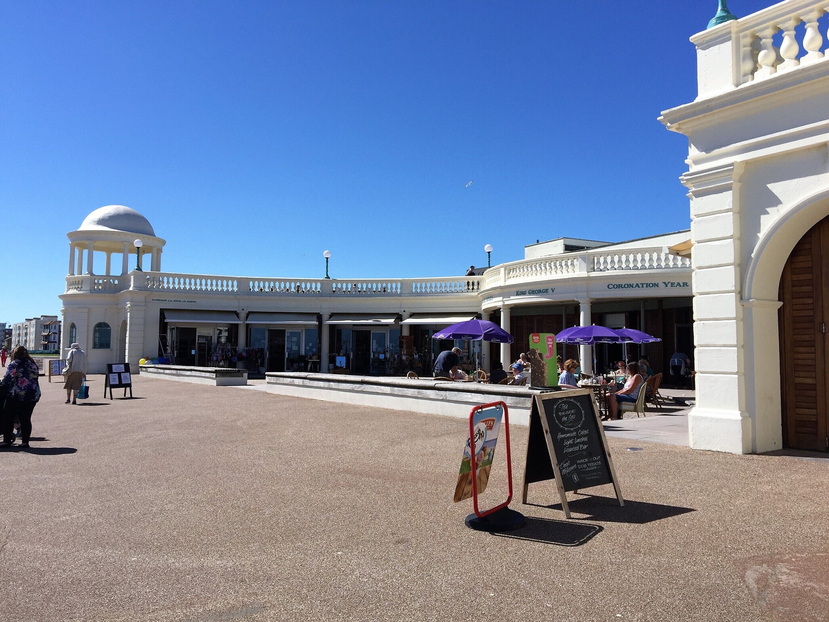 A public square with a white building featuring a dome and columns, outdoor seating with purple umbrellas, and a signboard with information.