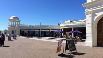 A public square with a white building featuring a dome and columns, outdoor seating with purple umbrellas, and a signboard with information.